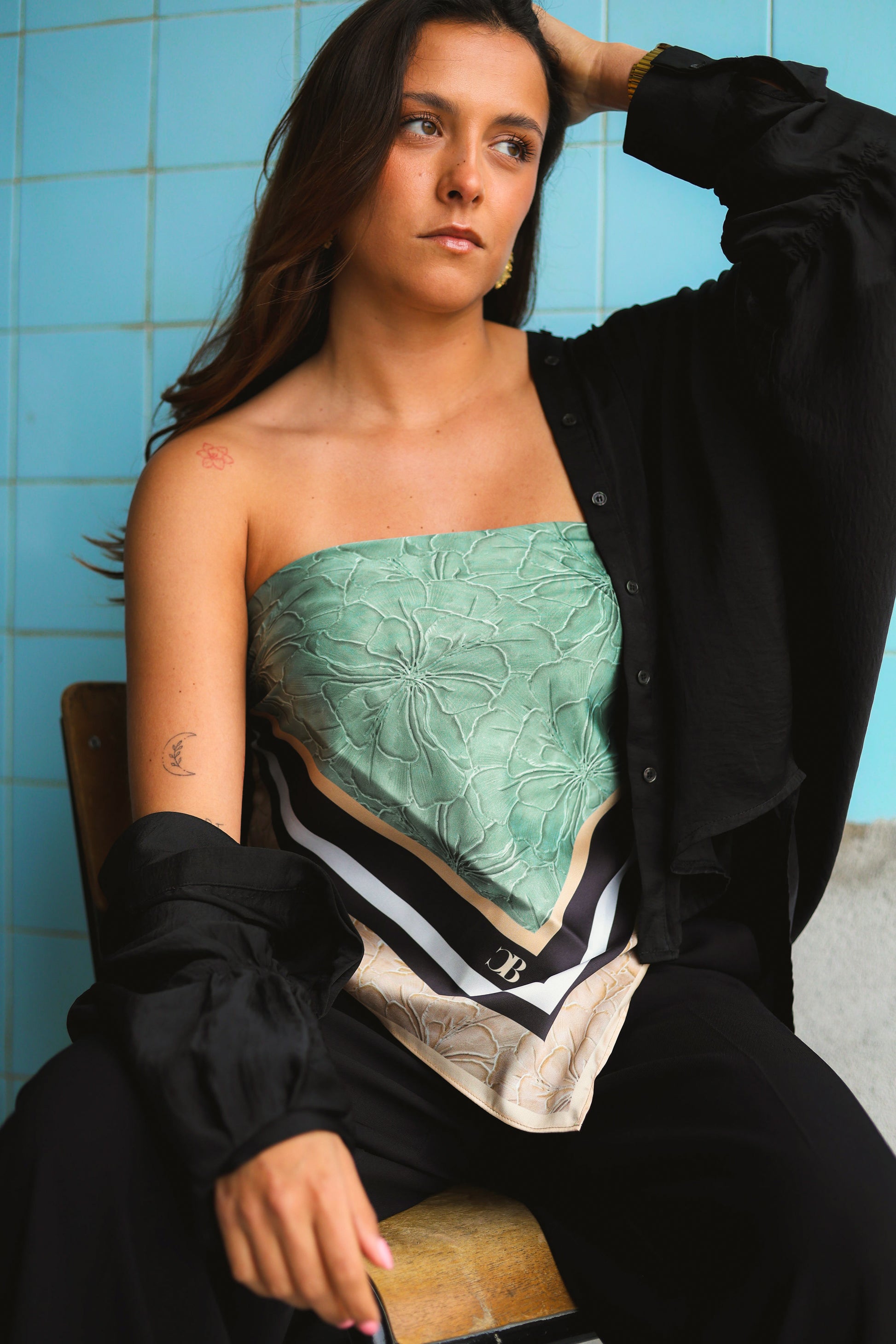 Woman wearing a green scarf with a black shirt over it, sitting against a blue tiled wall.
