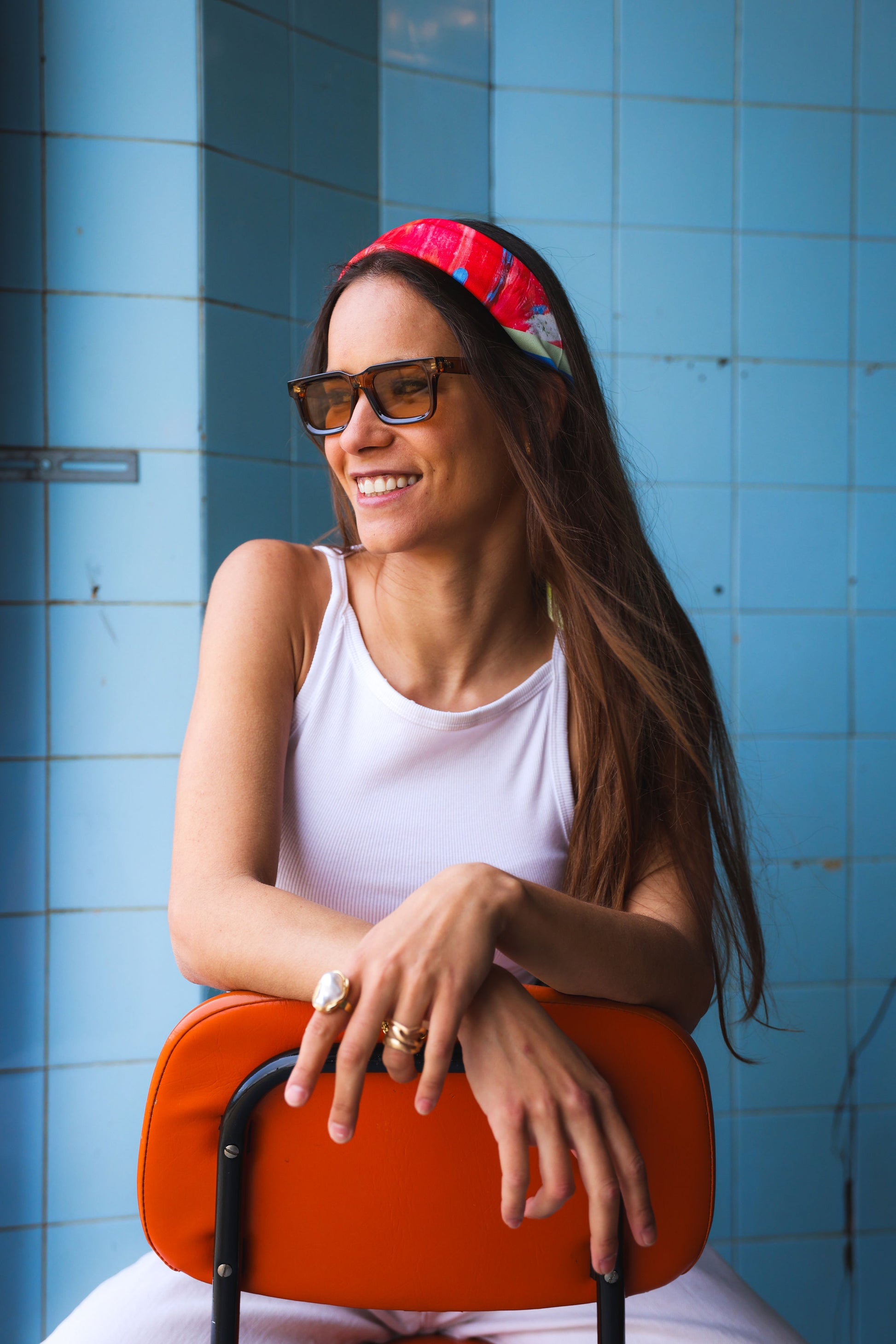 Woman with sunglasses and a colorful scarf sitting against a blue tiled wall.